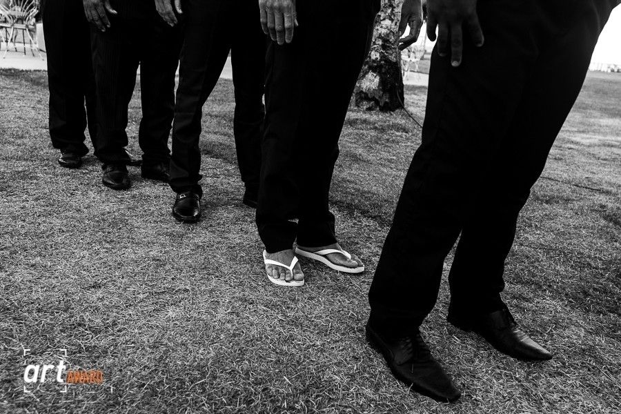 Black and white photo of men in dress shoes and one wearing flip flops standing on grass.