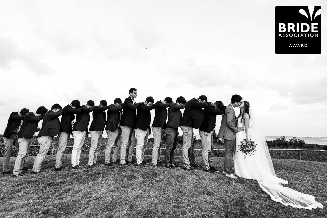 Black and white wedding photo of bride and groom kissing while groomsmen line up on a grassy hill.