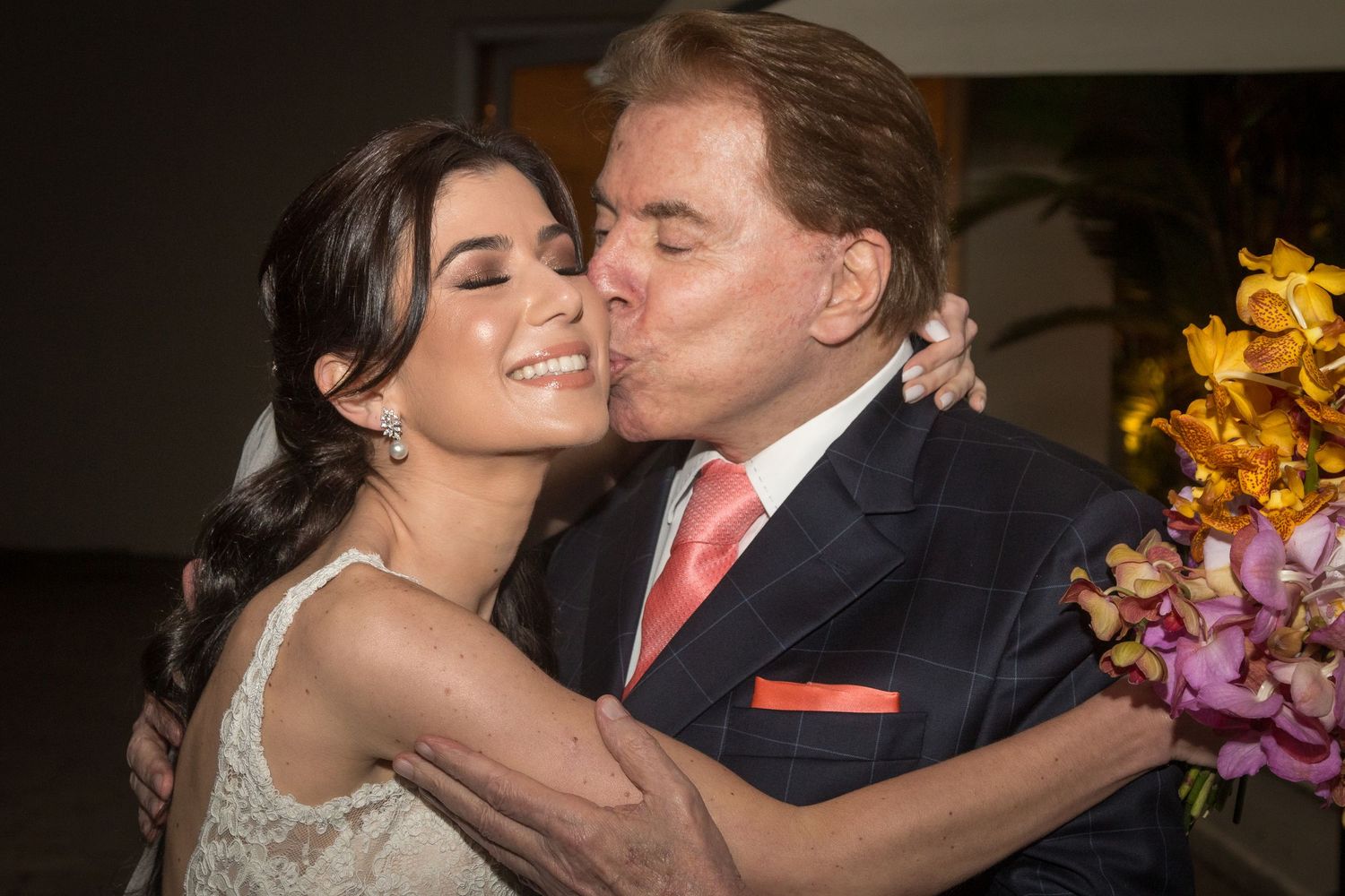 Elderly man in pink blazer kissing a young bride in white dress holding colorful flowers at a wedding celebration.