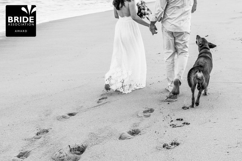 Black and white beach wedding photo of couple walking with dog, leaving footprints in sand, Bride Association Award winner.