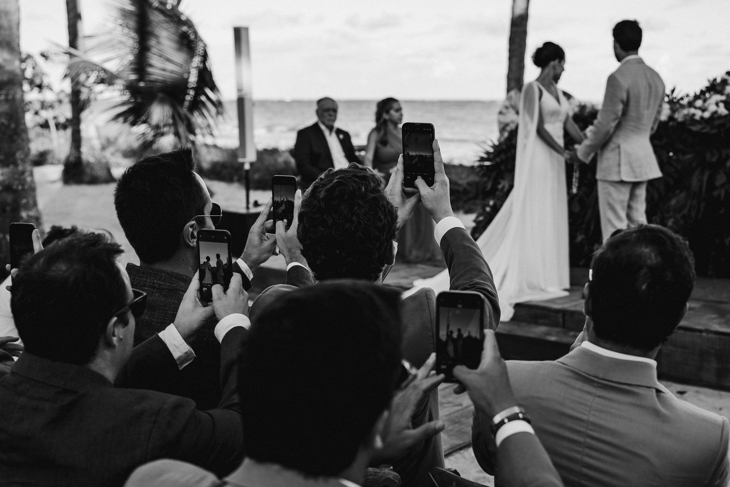 Black and white photo of wedding guests holding up phones to capture the beachside ceremony.
