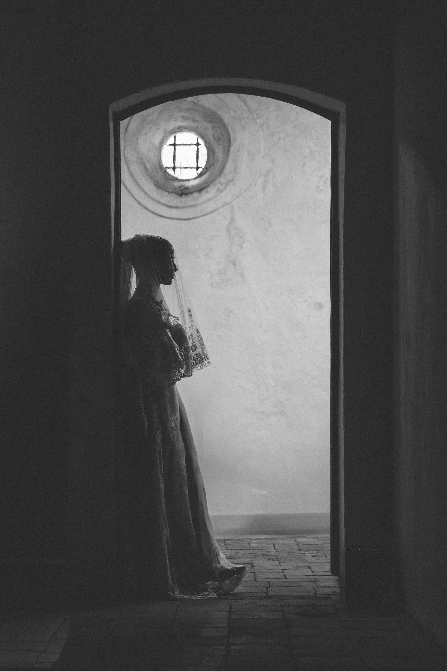 Black and white silhouette of a bride in a wedding dress and veil standing in an arched stone doorway.