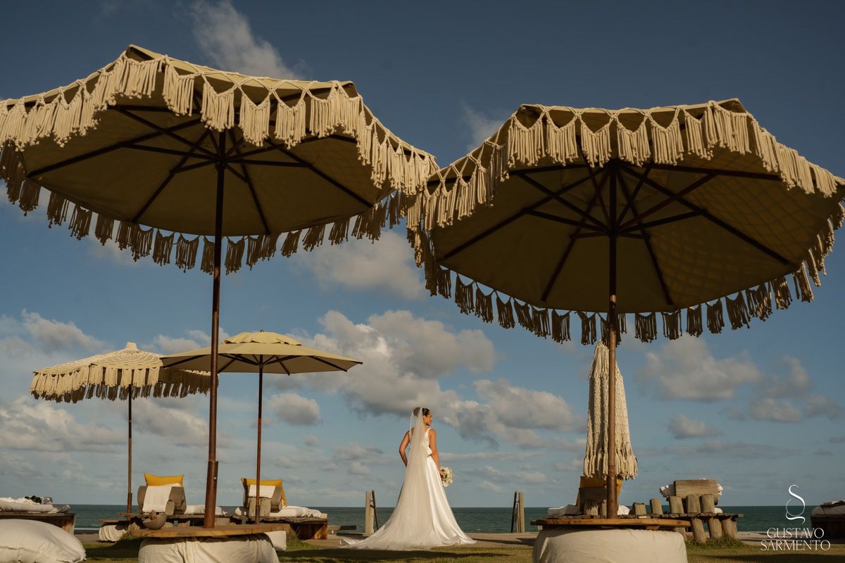 Bride in white gown standing among elegant fringed beach umbrellas with ocean backdrop.