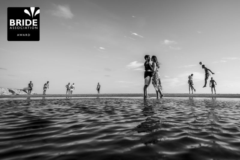 Black and white wedding photo of couple embracing in shallow water surrounded by silhouetted figures along the horizon.