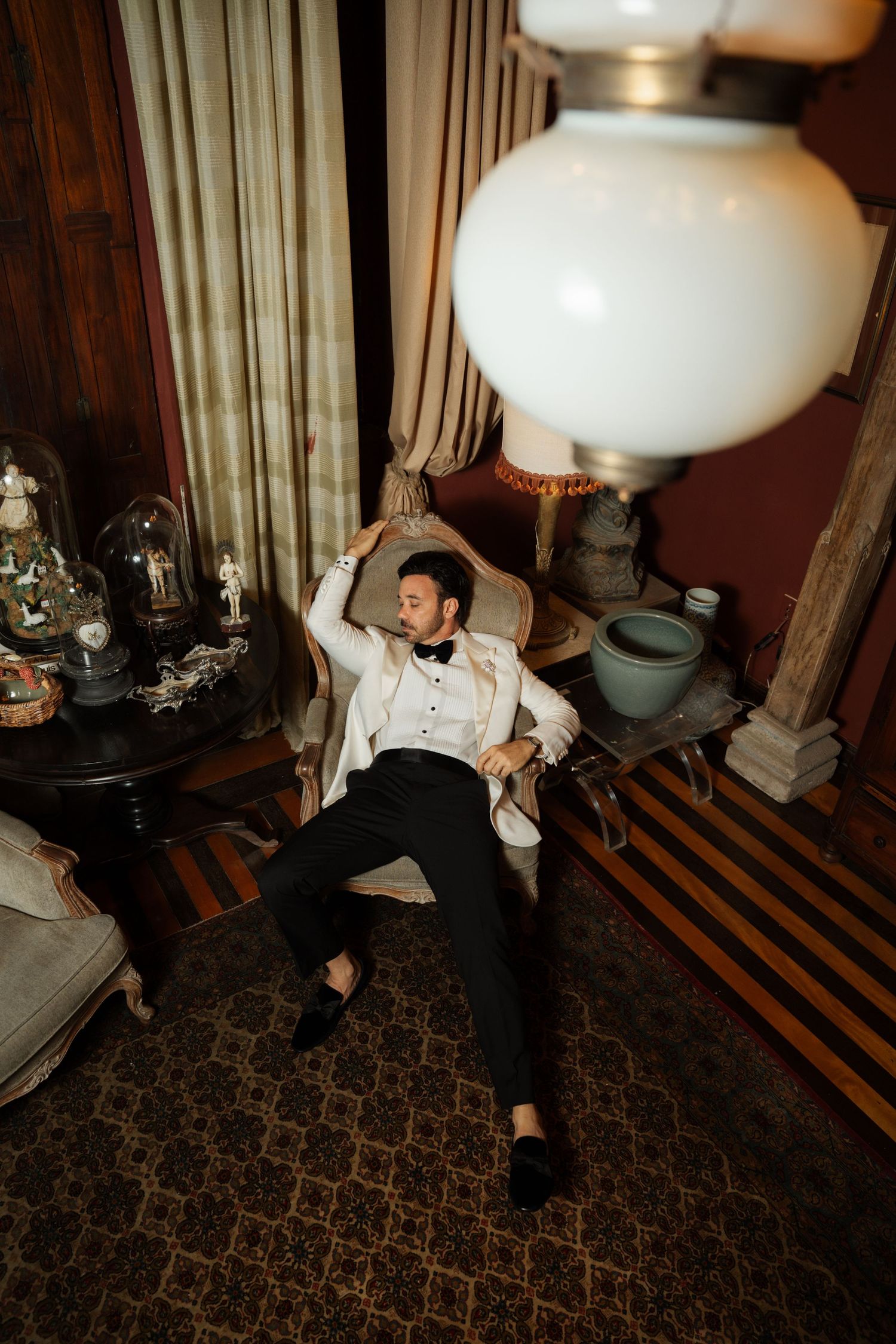 A groom in a white tuxedo jacket and black trousers relaxes in a vintage chair in an elegantly decorated room.