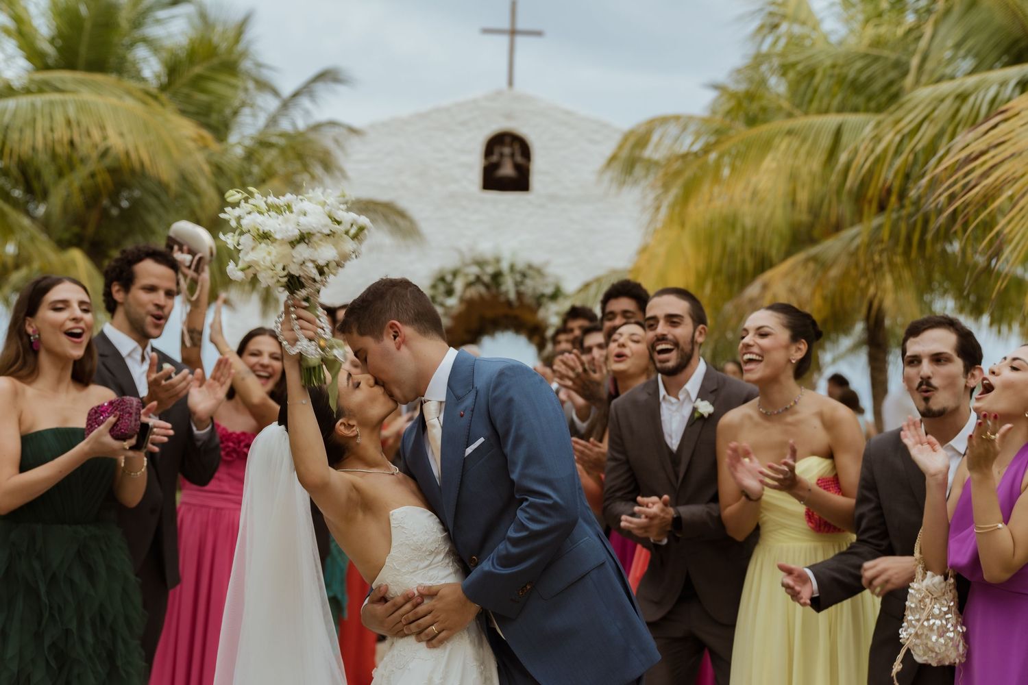 Bride and groom share a kiss surrounded by cheering wedding guests near a tropical church with palm trees.