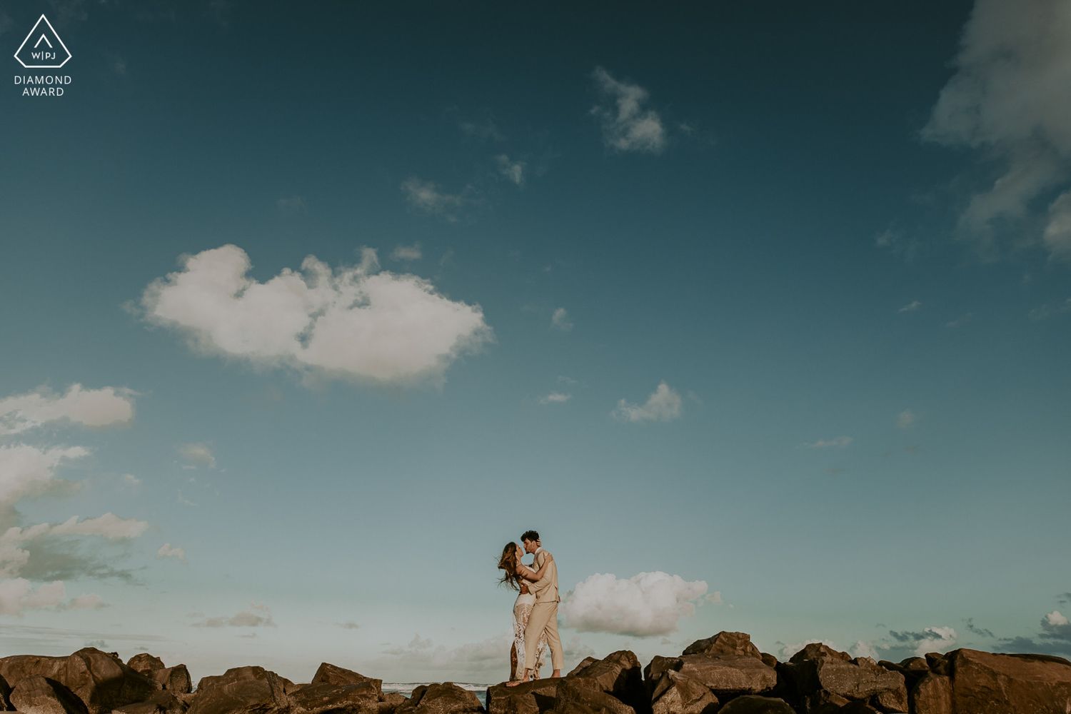 Couple embracing on rocky jetty under vast blue sky with scattered clouds during romantic outdoor engagement photo session.