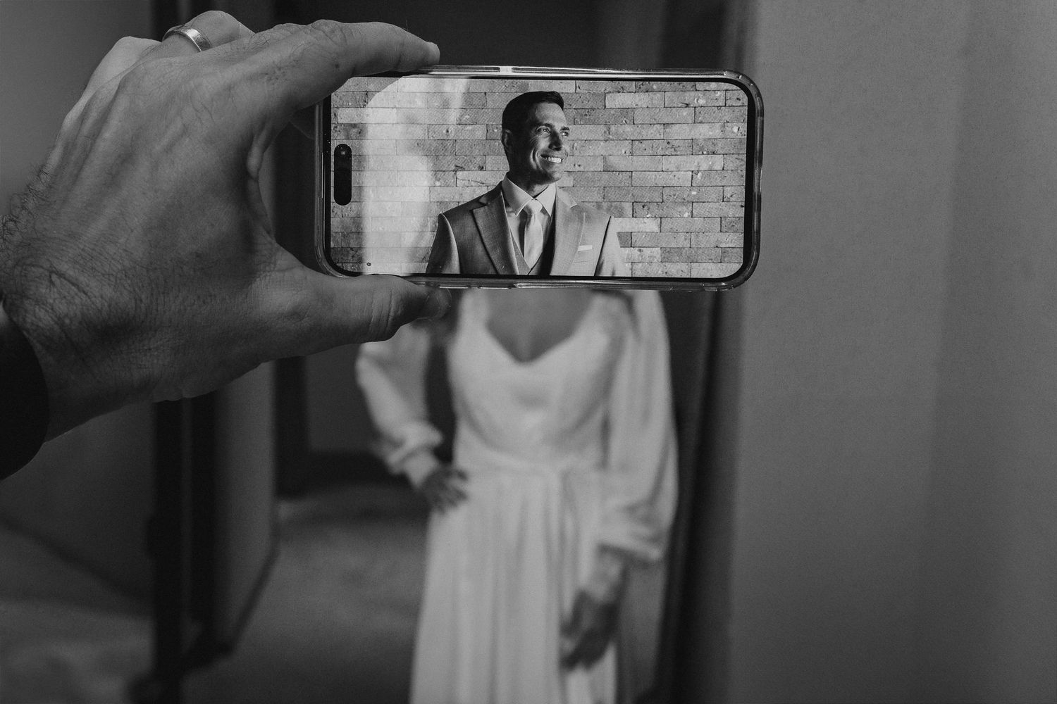 Black and white photo of a groom reflected in a phone screen held up in front of a bride in a wedding dress.