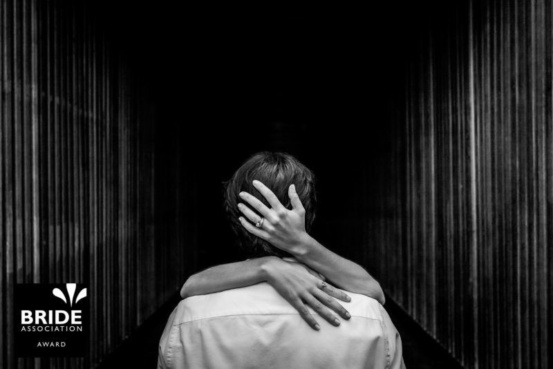 Black and white photo of a couple embracing from behind in a dramatic columned hallway at a wedding.