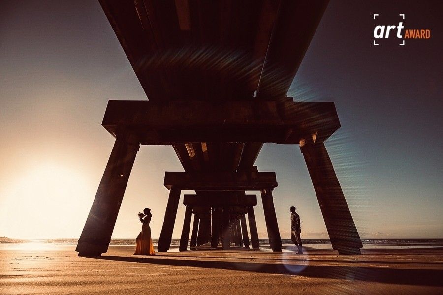 Silhouettes of two people standing beneath a wooden pier at sunset on a sandy beach, with golden light filtering through the structure.
