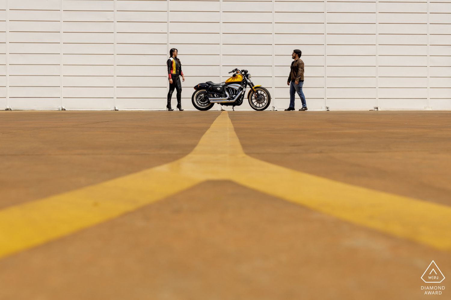 Two people stand apart beside a yellow motorcycle on a concrete lot with bold yellow lines and a white corrugated wall backdrop.