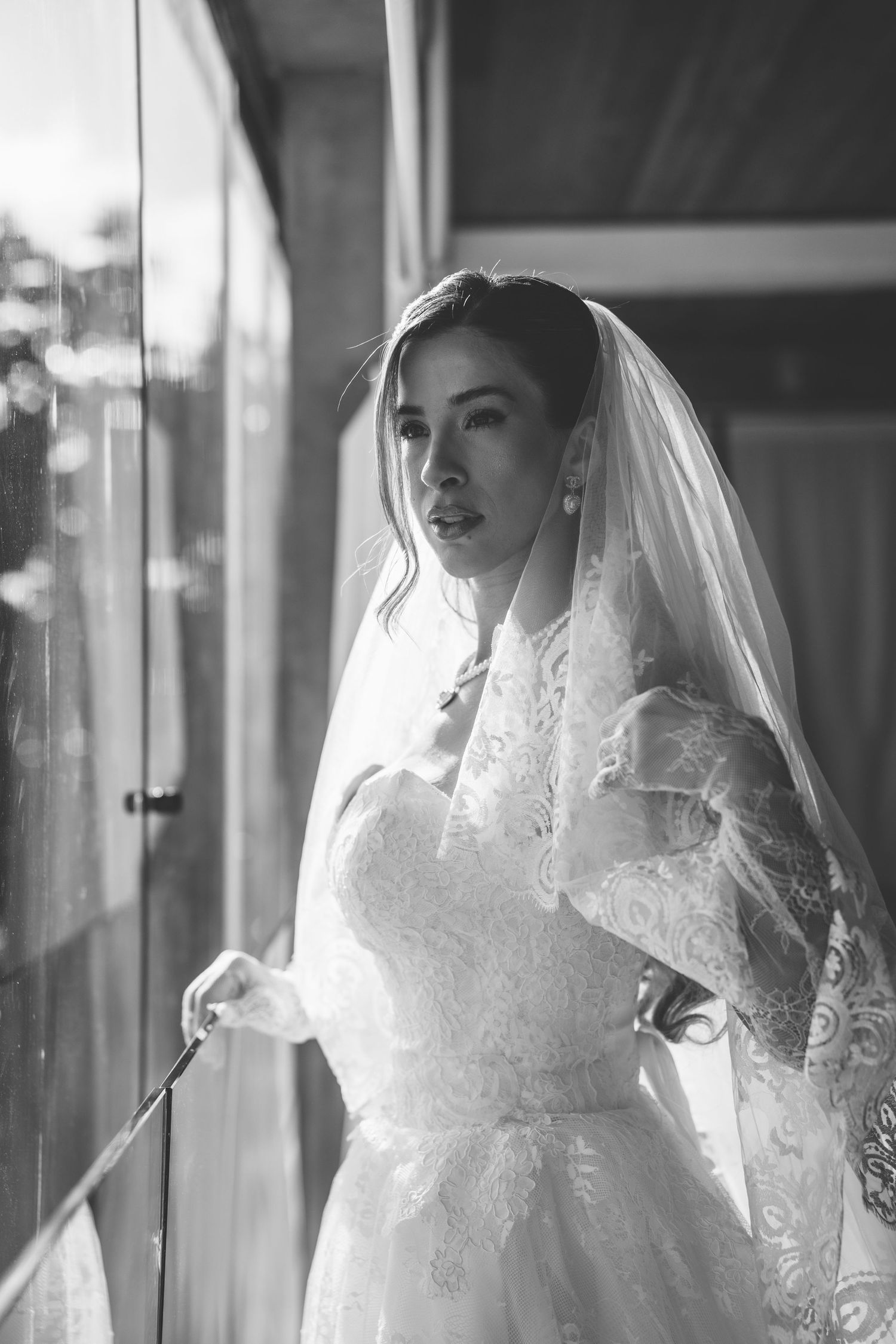 Black and white photo of a bride in lace wedding dress and veil standing by a window in soft natural light.