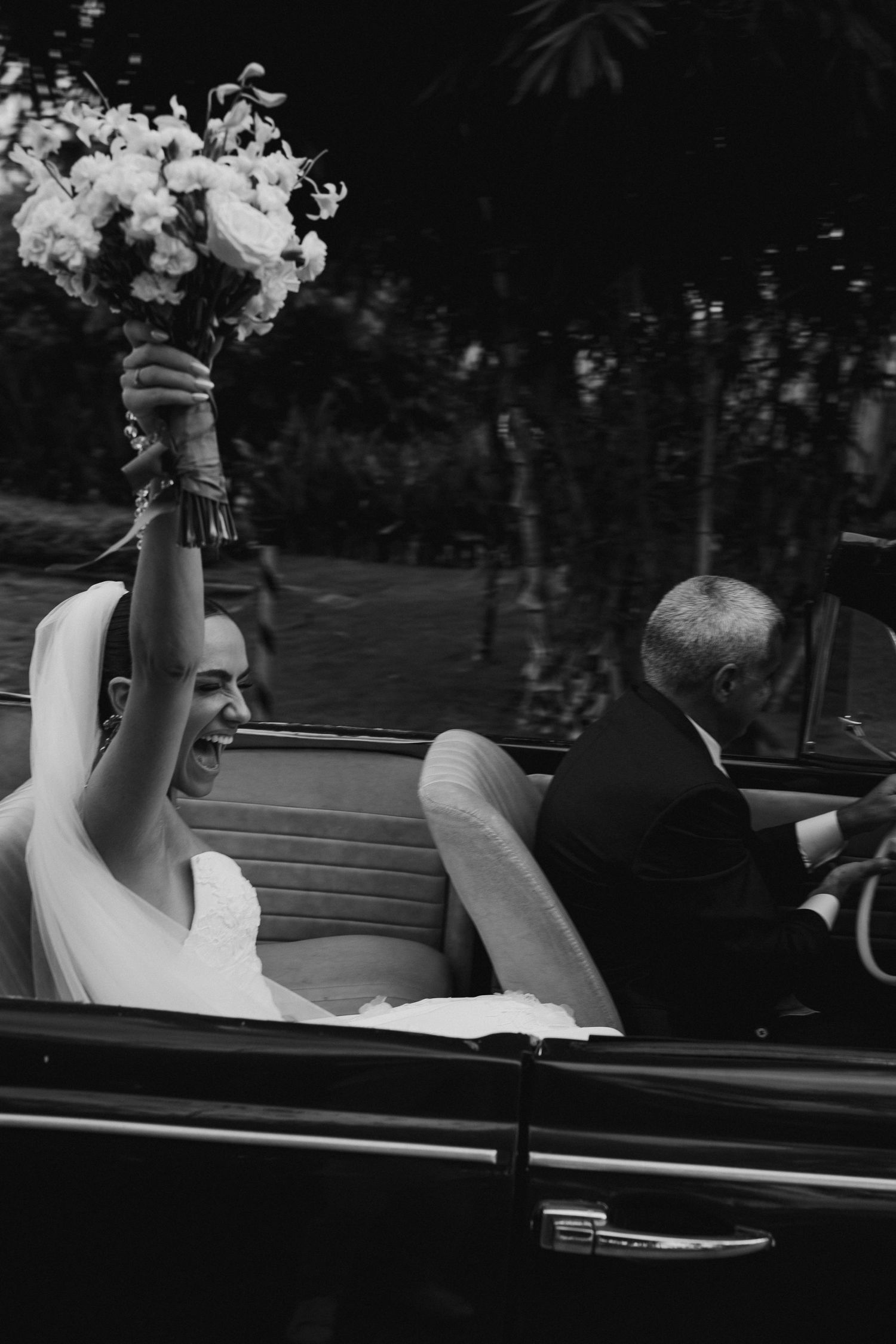 Joyful bride raises bouquet while riding in a classic convertible car on her wedding day.