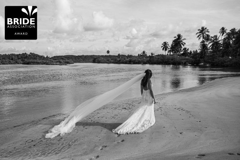 Bride in flowing gown and veil walks along tropical beach in stunning black and white wedding photo.