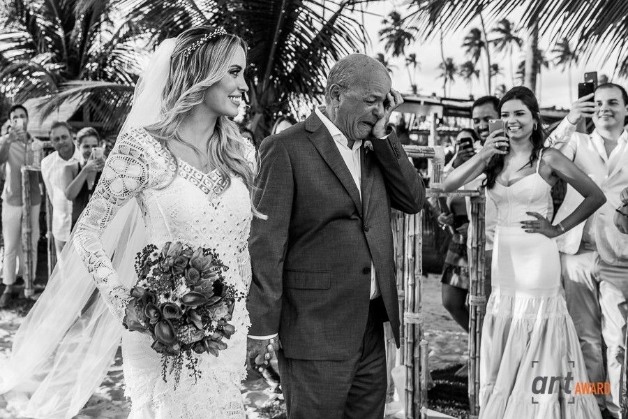 Black and white beach wedding photo of bride in lace gown holding bouquet, escorted by older man, palm trees behind.