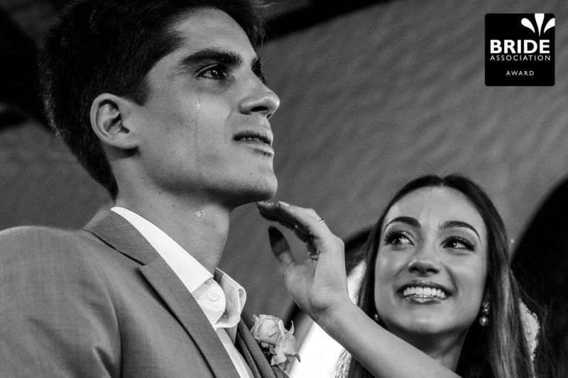 Black and white wedding photo of groom in suit with boutonniere as bride smiles beside him.