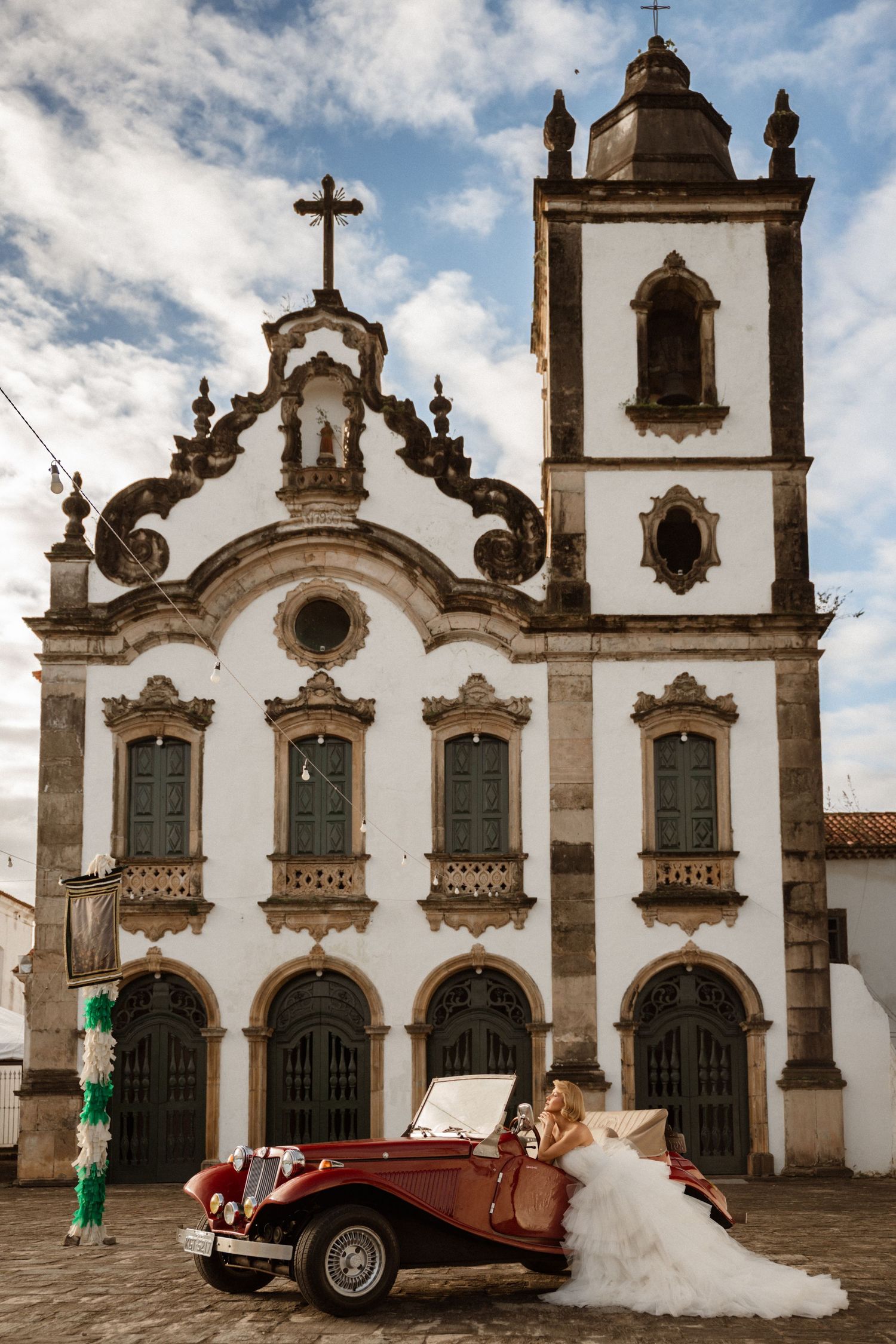 Bride in white gown poses beside vintage red convertible in front of ornate white Baroque church under blue sky.
