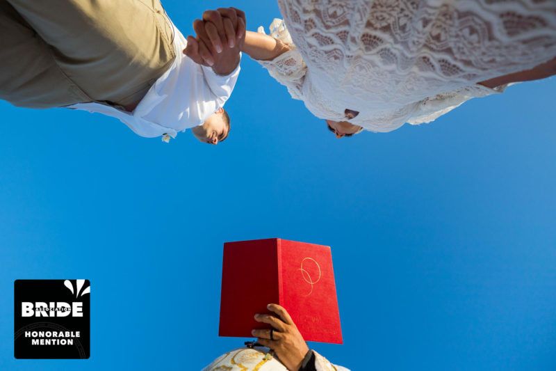 Creative low-angle wedding photo showing couple holding hands above officiant reading red book against blue sky.