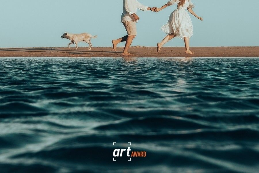 Couple in white holding hands running barefoot on beach with dog, low angle water view, Art Award logo.