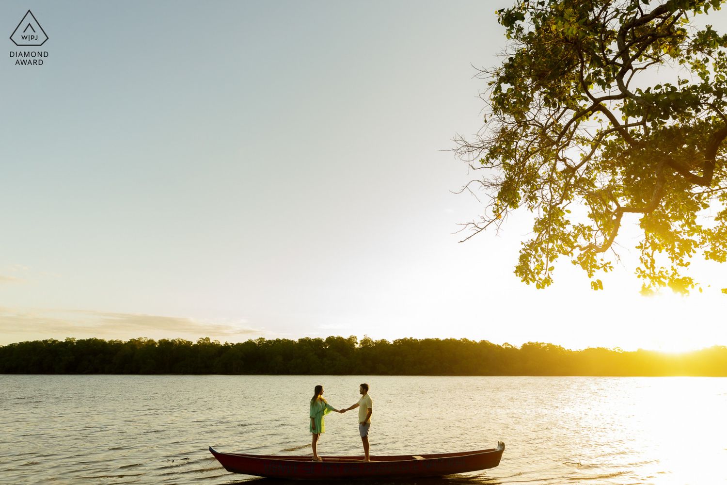 Couple holding hands standing in a red canoe on a serene lake at golden sunset with lush trees in background.
