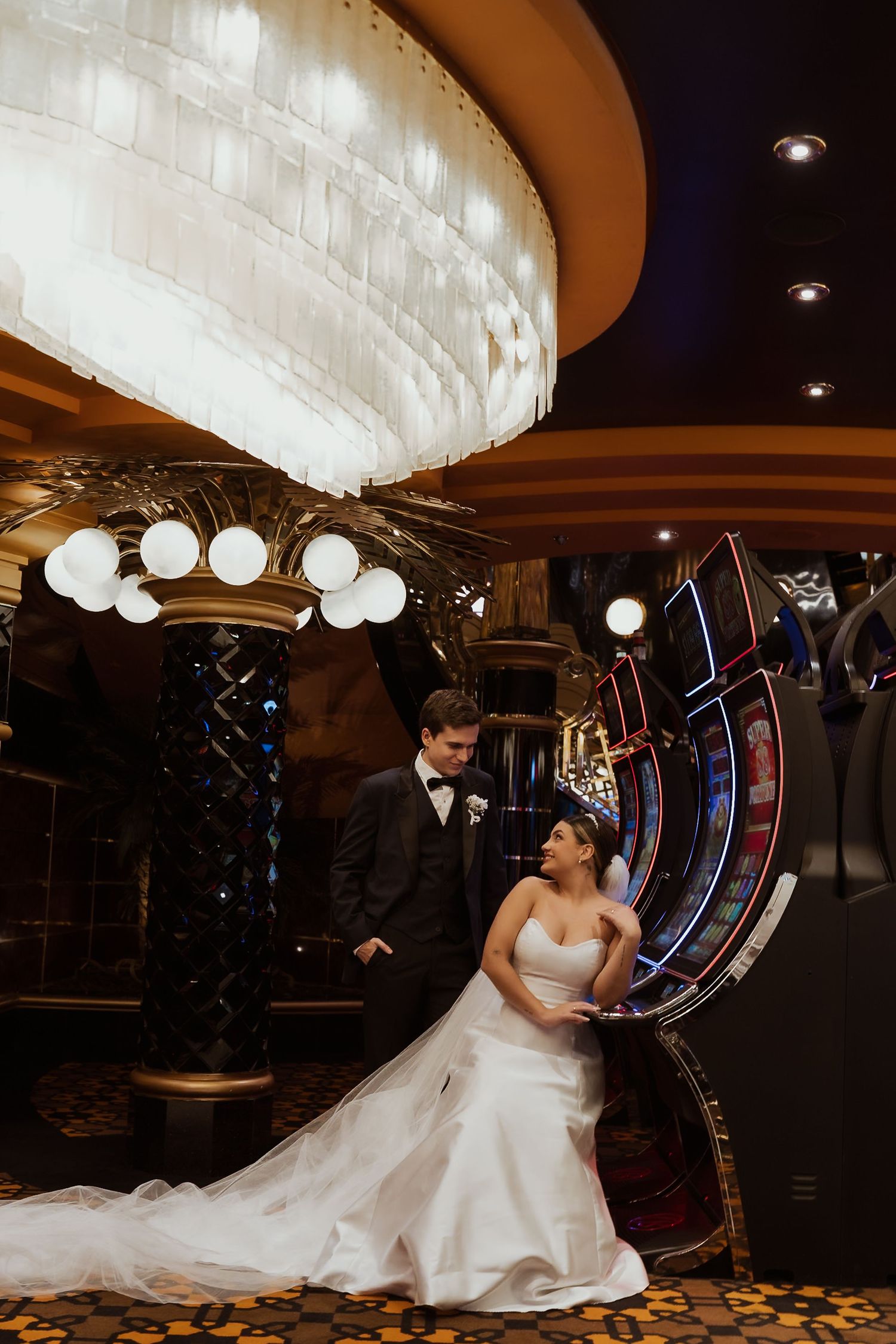 Bride and groom pose elegantly beside a roulette wheel under a grand chandelier in a luxurious casino venue.