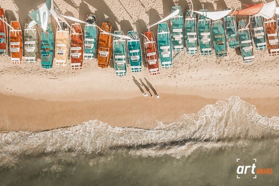 Aerial view of colorful boats lined up on a sandy beach with gentle ocean waves.