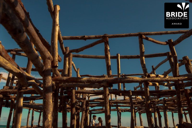 Rustic wooden scaffolding structure on a tropical beach with clear blue sky, featuring a Bride Association Award logo.