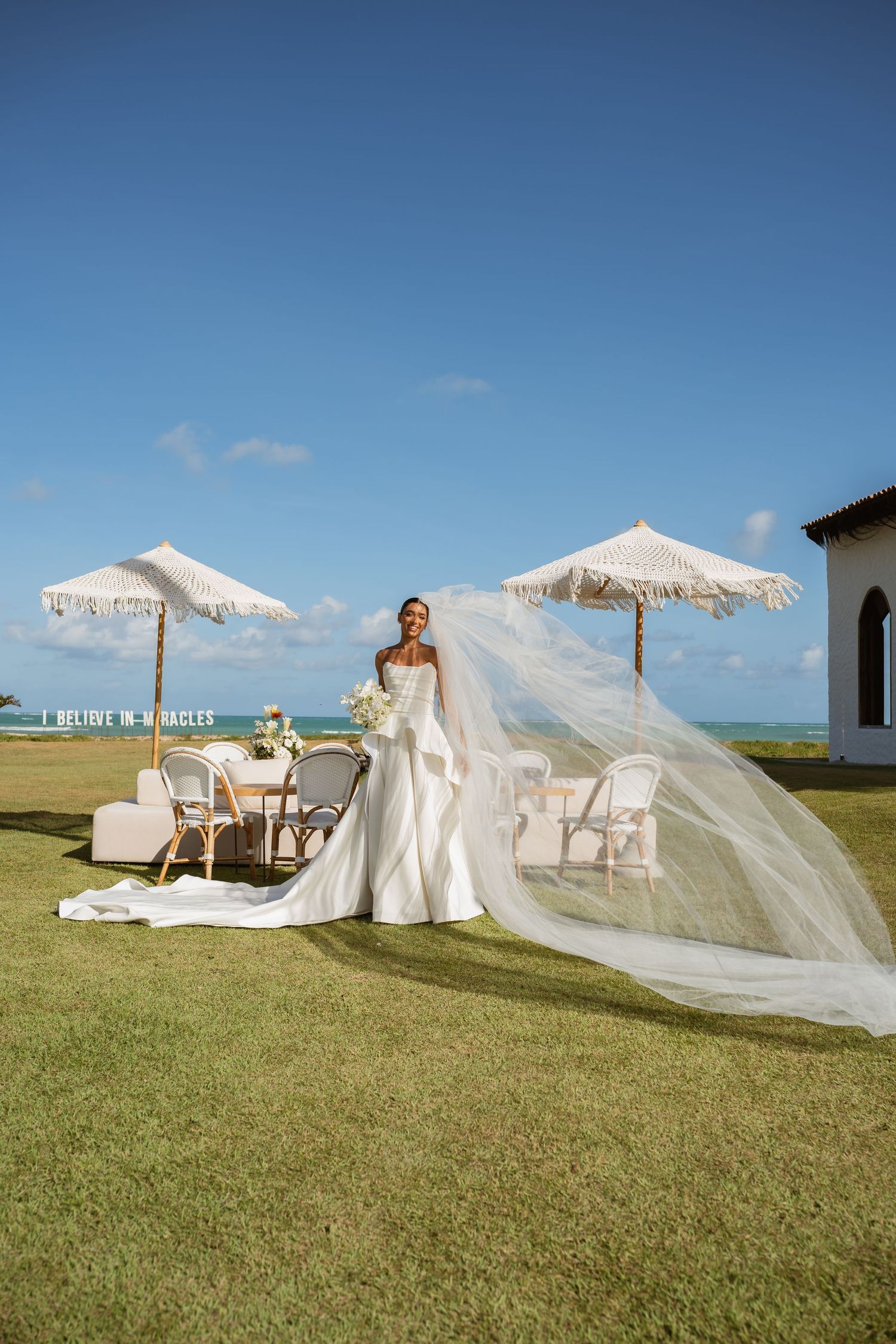 Bride in flowing white gown with veil blowing in wind beside fringed umbrellas by the sea.