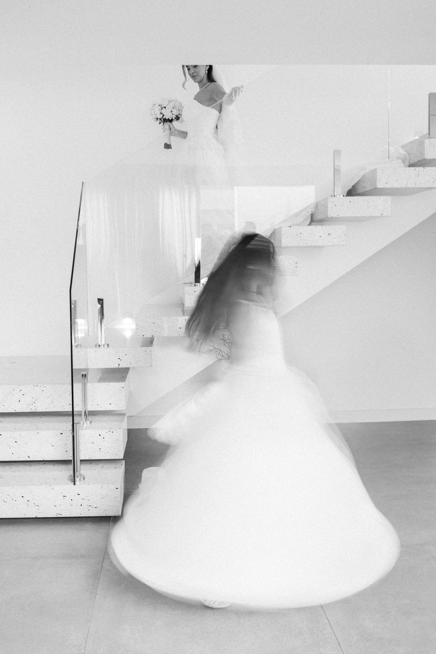Black and white photo of a bride in a white gown spinning gracefully near a modern staircase, creating a dreamy motion blur effect.