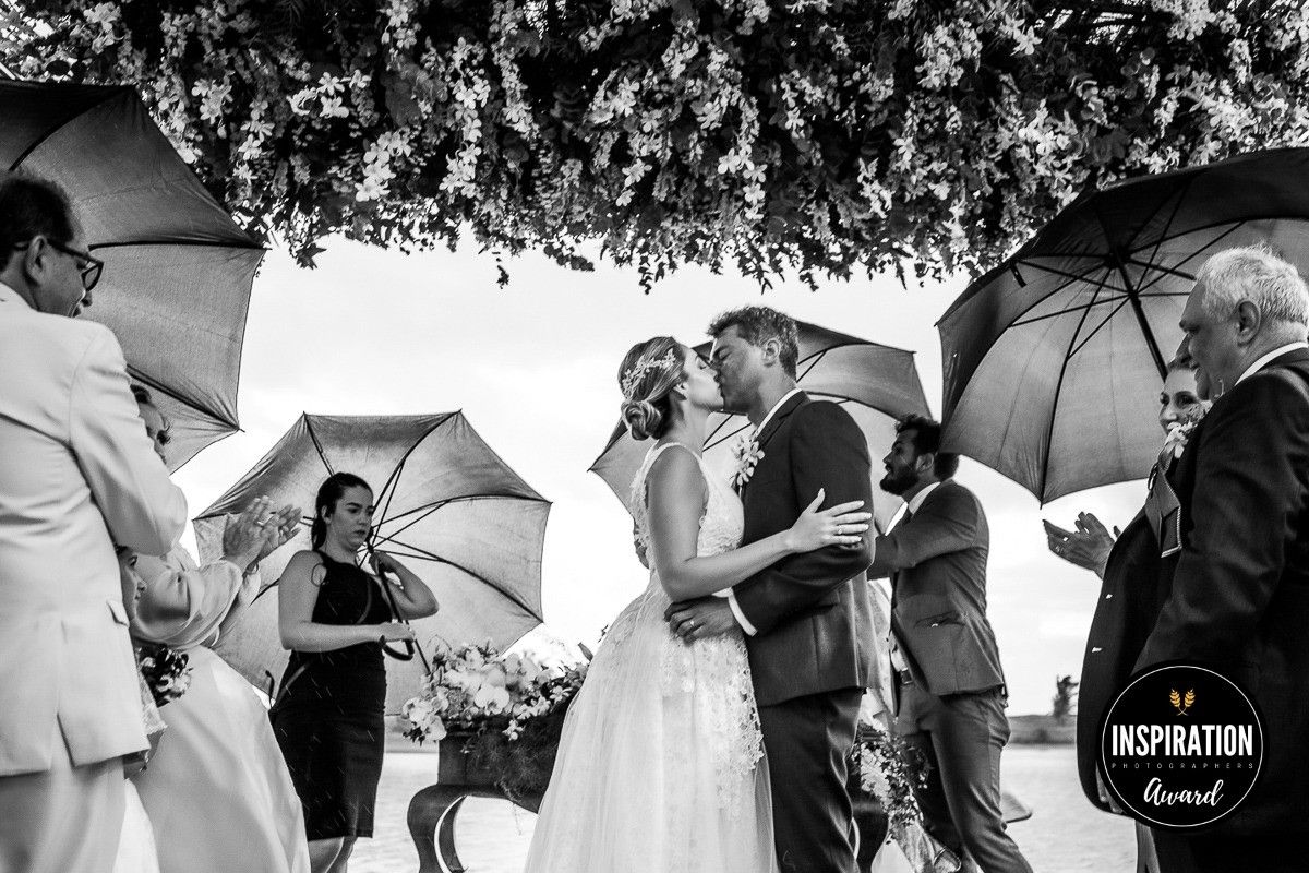 Black and white wedding kiss under umbrellas in rain, bride in white gown, groom in suit, guests surrounding couple.