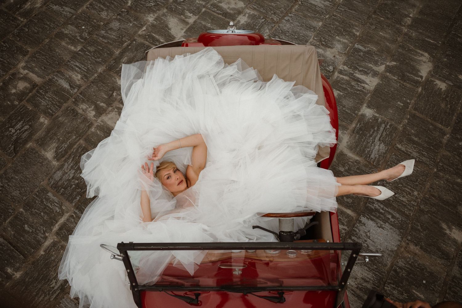 Bride in white gown lying elegantly in a red vintage suitcase, aerial view on wooden floor.
