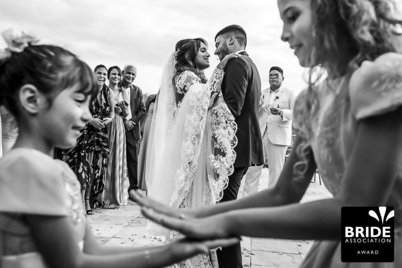 Black and white wedding ceremony photo with bride and groom exchanging vows outdoors, surrounded by guests and flower girls.