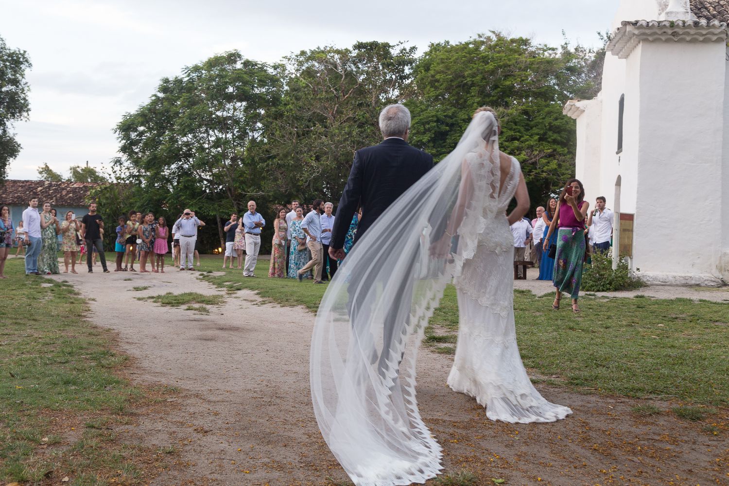Bride in white gown with long lace veil walks with escort toward outdoor wedding ceremony guests.