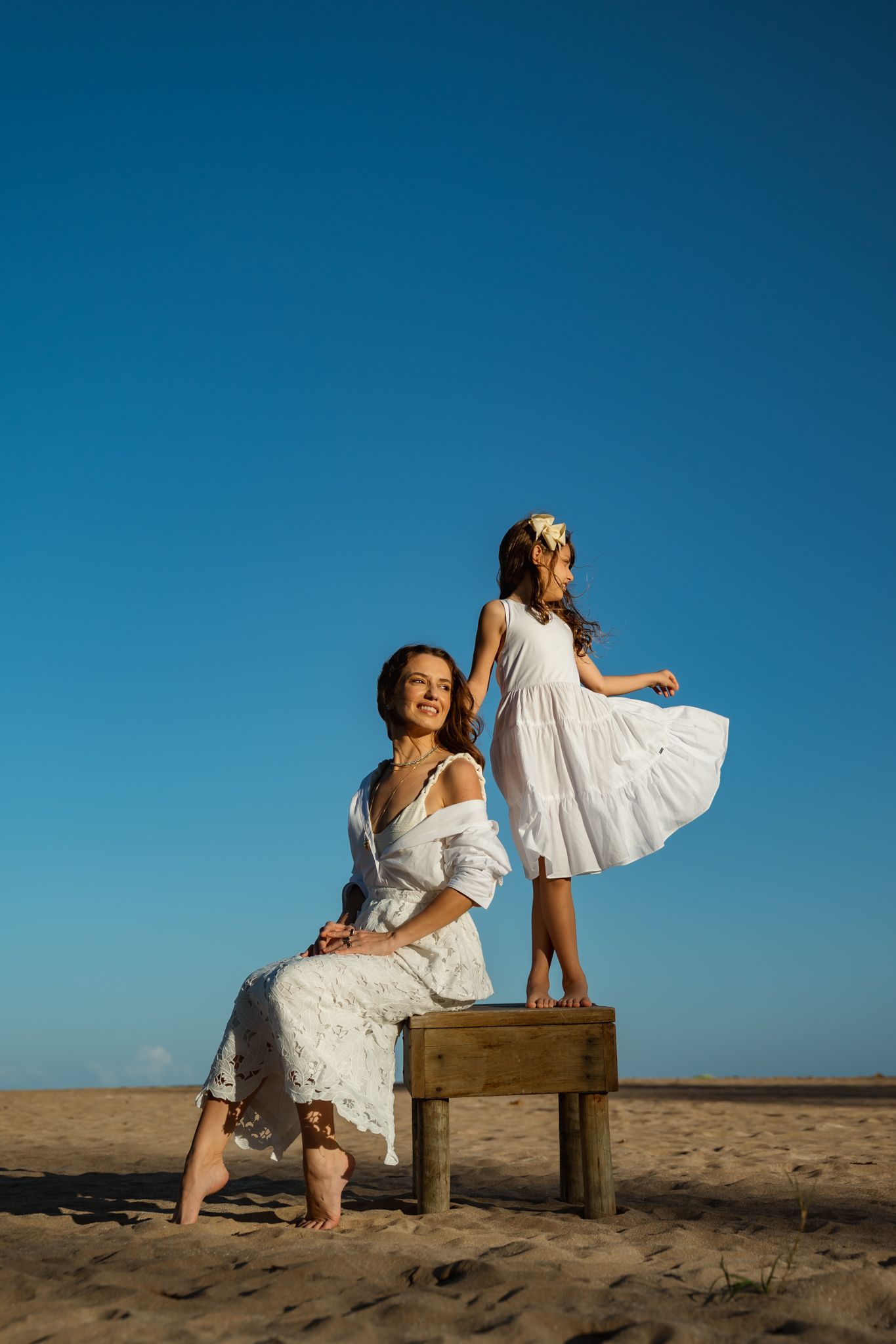 Mother and daughter in white dresses on sandy beach, girl standing on wooden bench under clear blue sky.