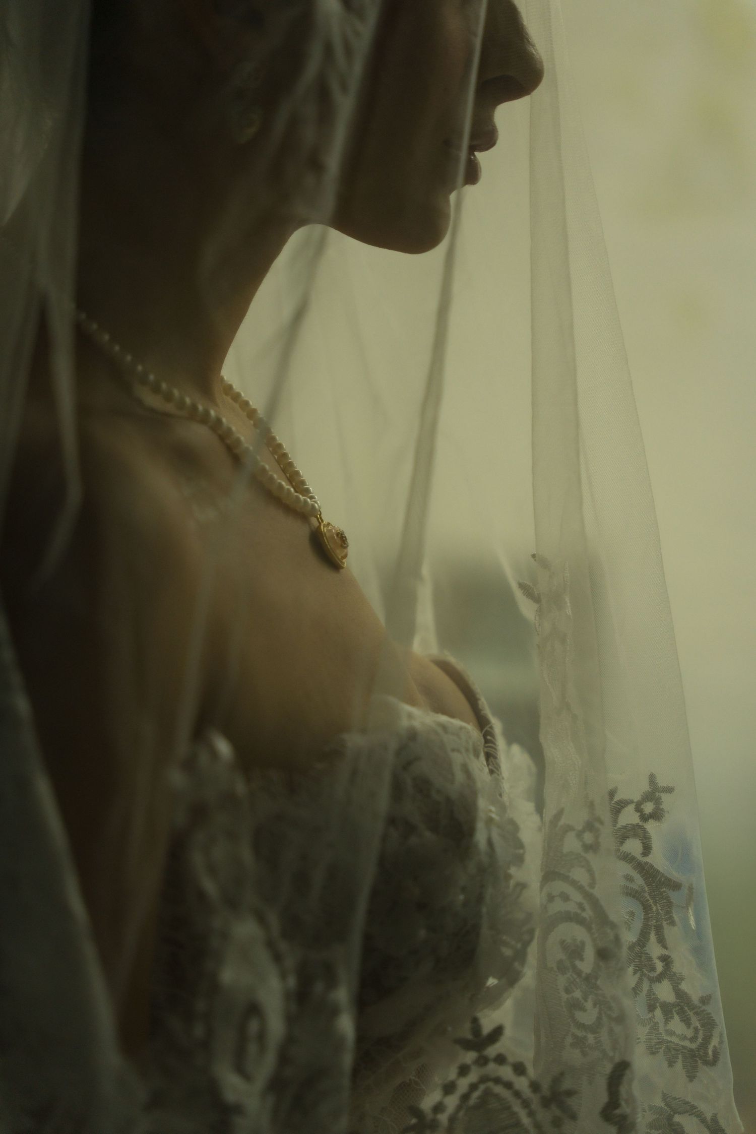 Close-up silhouette of a bride in lace wedding dress wearing pearl necklace and veil by a window.