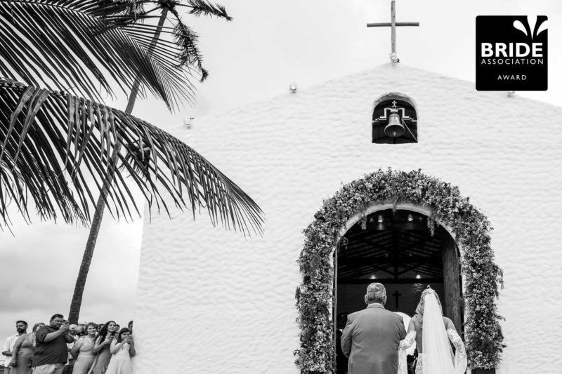 Black and white photo of a bride entering a floral-adorned chapel with a palm tree.