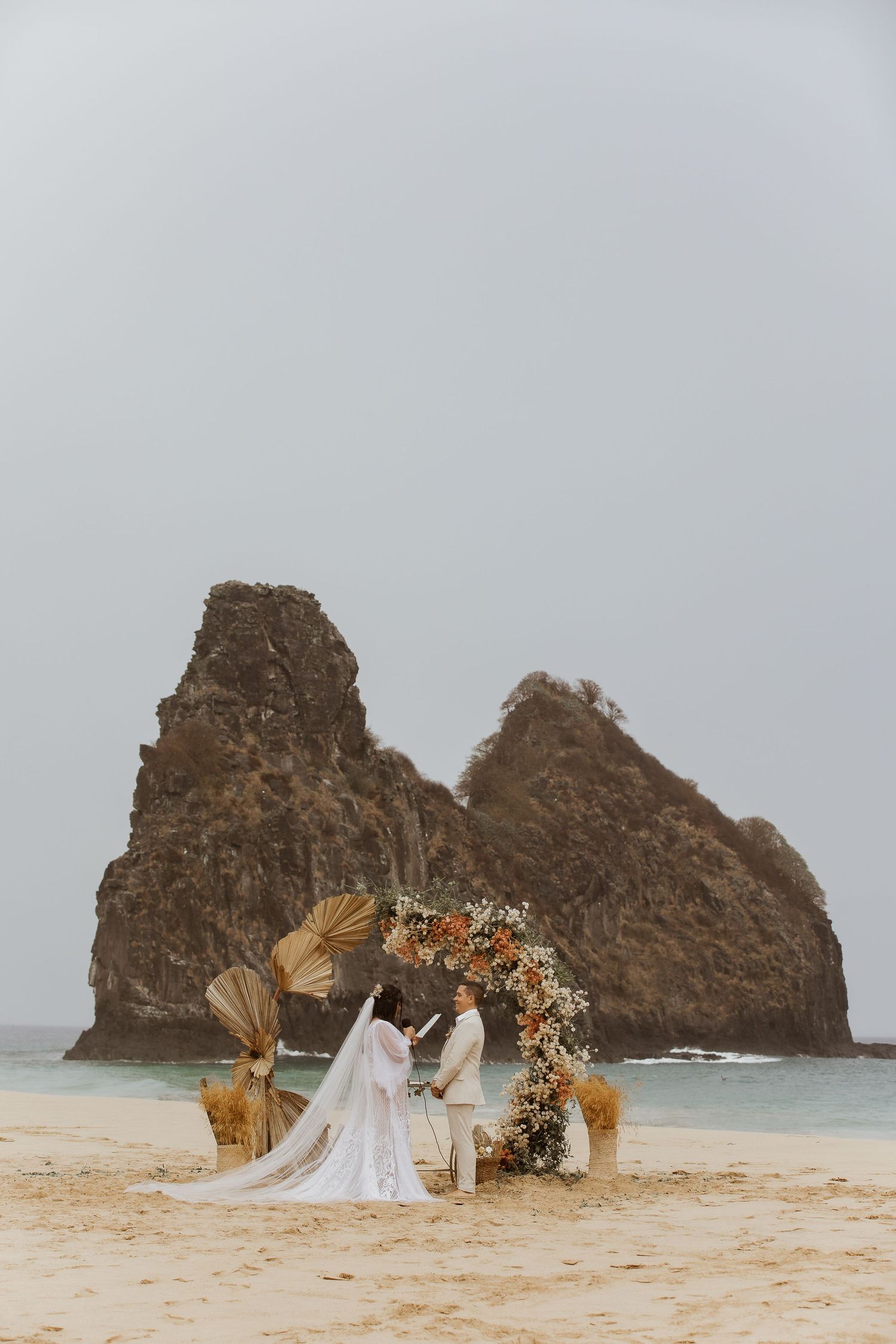 Couple exchanging vows at a boho beach wedding ceremony with floral arch and dramatic rock formations in the background.
