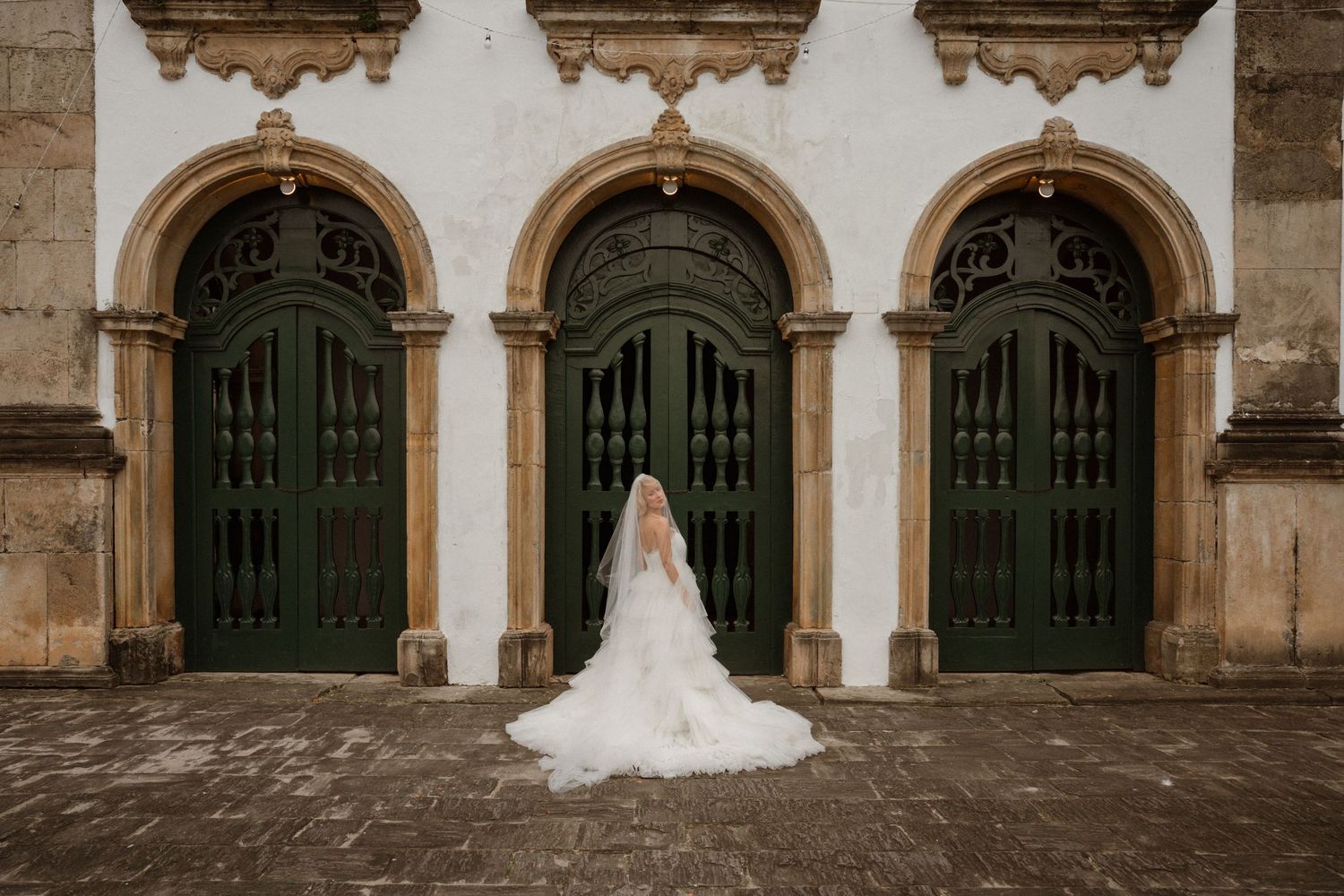 Bride in white gown and veil stands before ornate green arched doors of historic stone building.