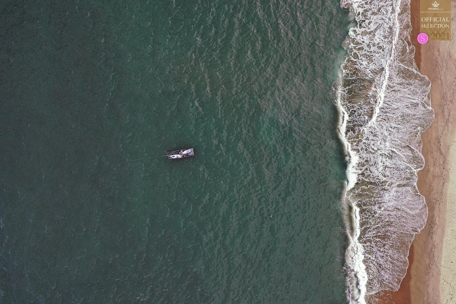 Aerial view of a surfer paddling in turquoise ocean waves near a sandy beach shoreline.