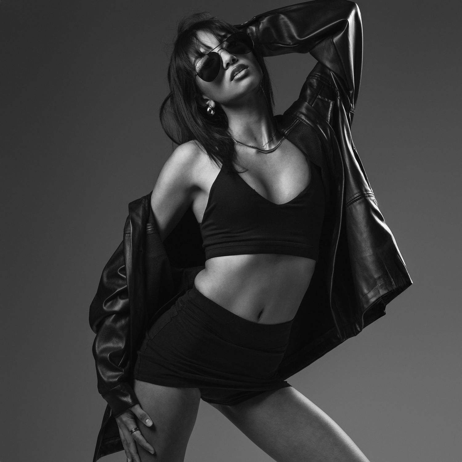 Black and white studio portrait of a woman in sunglasses, crop top, shorts and an oversized leather jacket striking a confident pose.