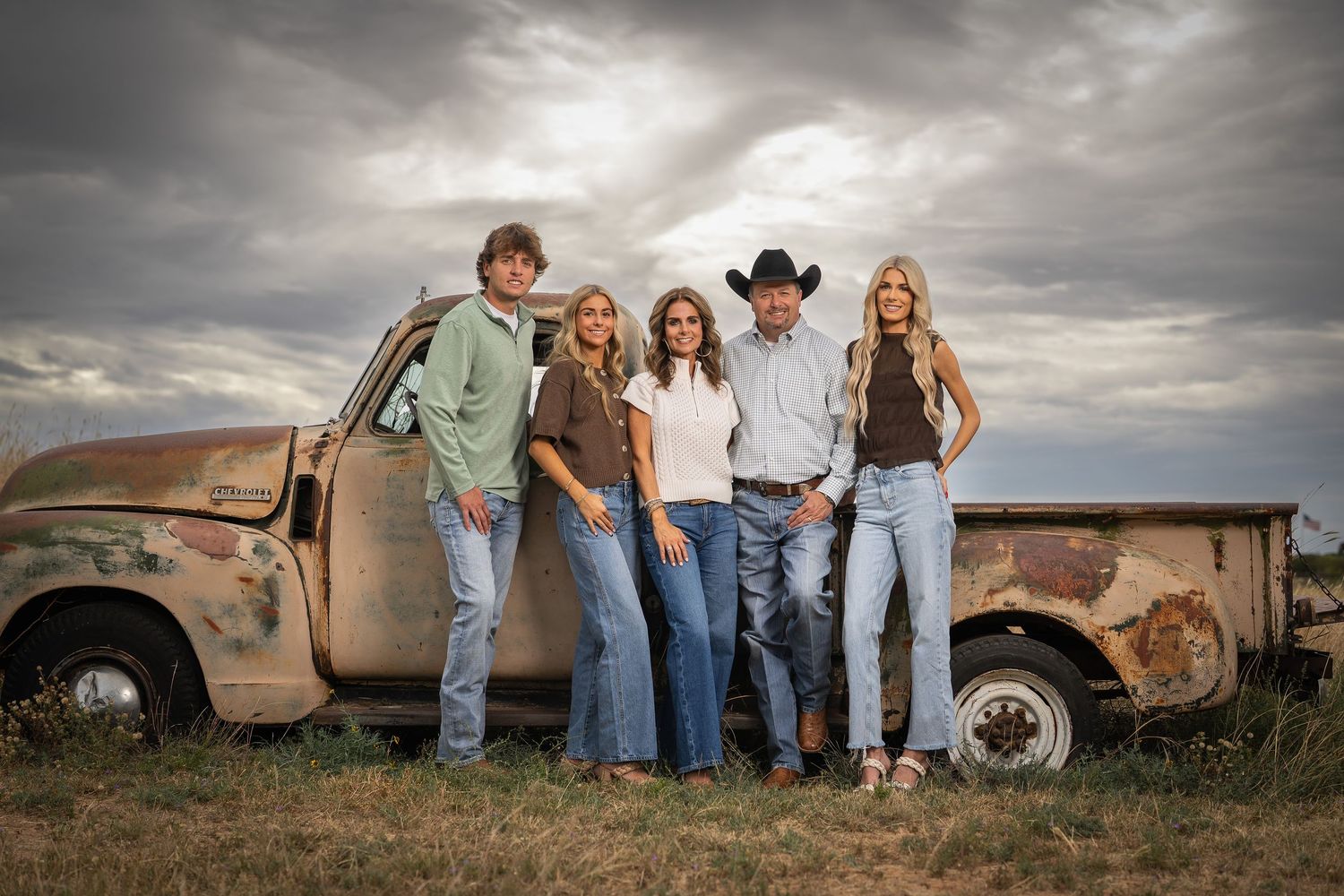 A group poses casually against a rustic vintage truck under dramatic cloudy skies in a country setting.