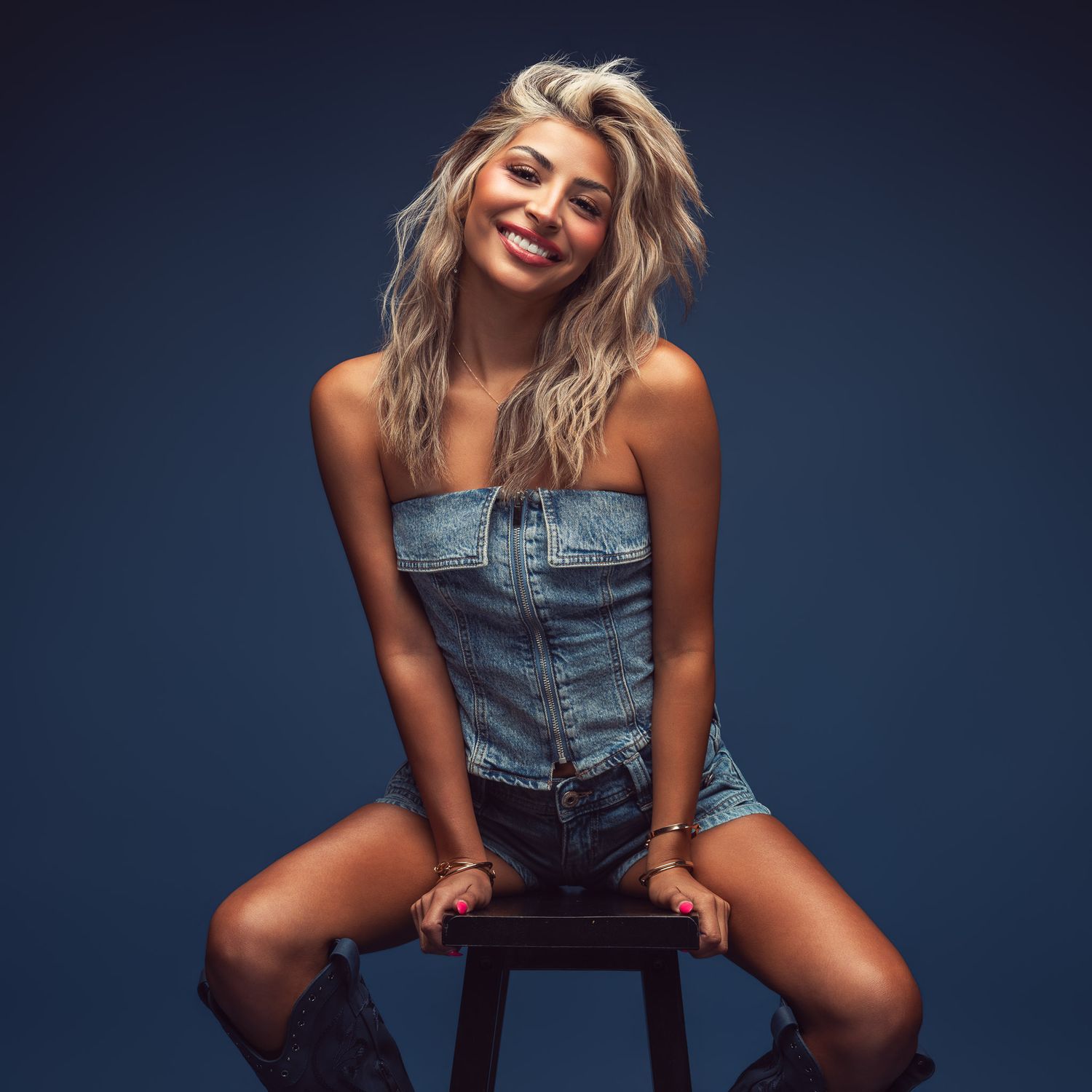 Smiling woman with wavy blonde hair in a denim strapless romper and black boots sits on a wooden stool against a dark blue studio backdrop.
