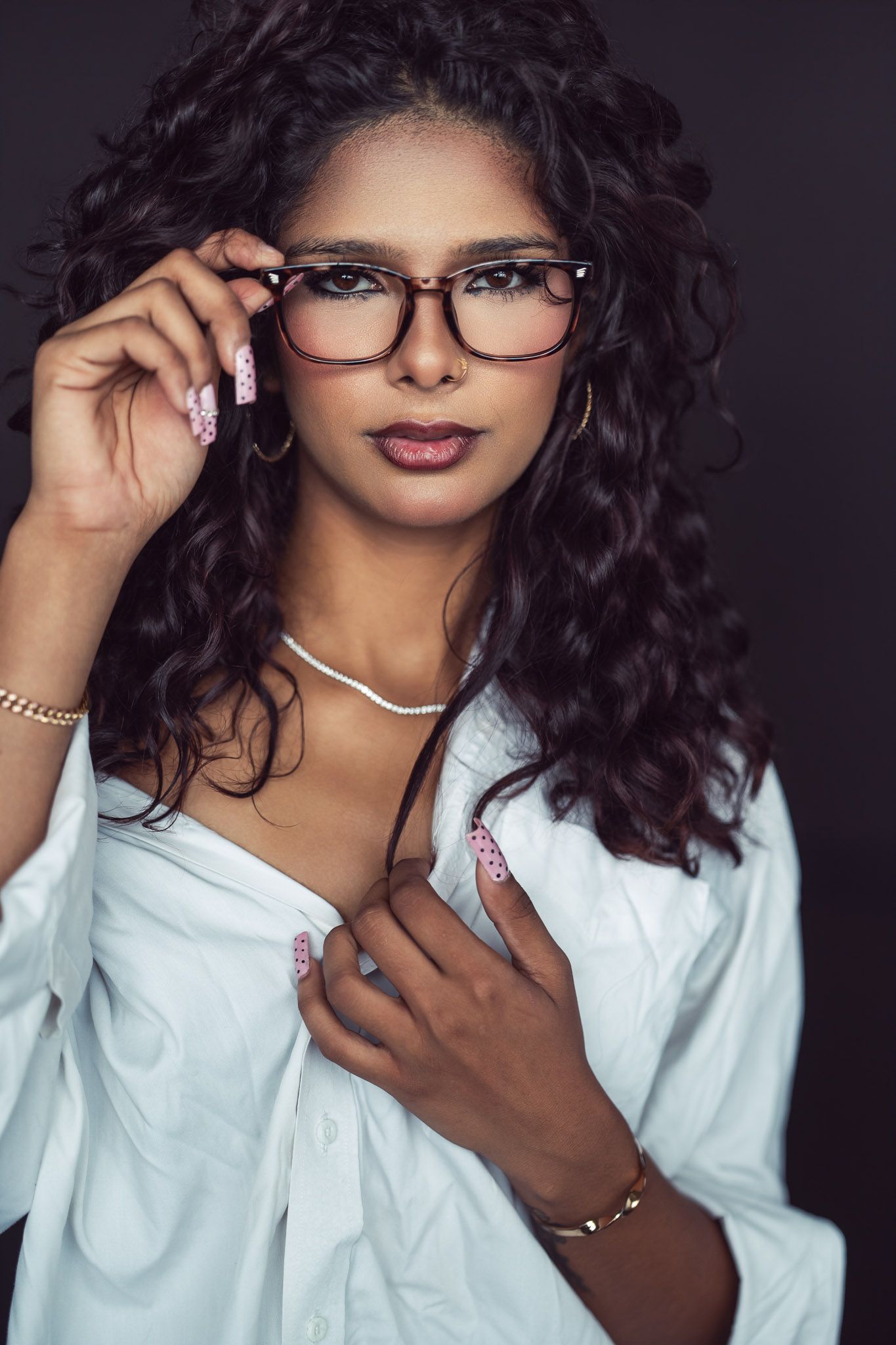 Woman with long curly dark hair adjusts tortoiseshell glasses, wearing a white button-up shirt, gold jewelry and pink nail art.