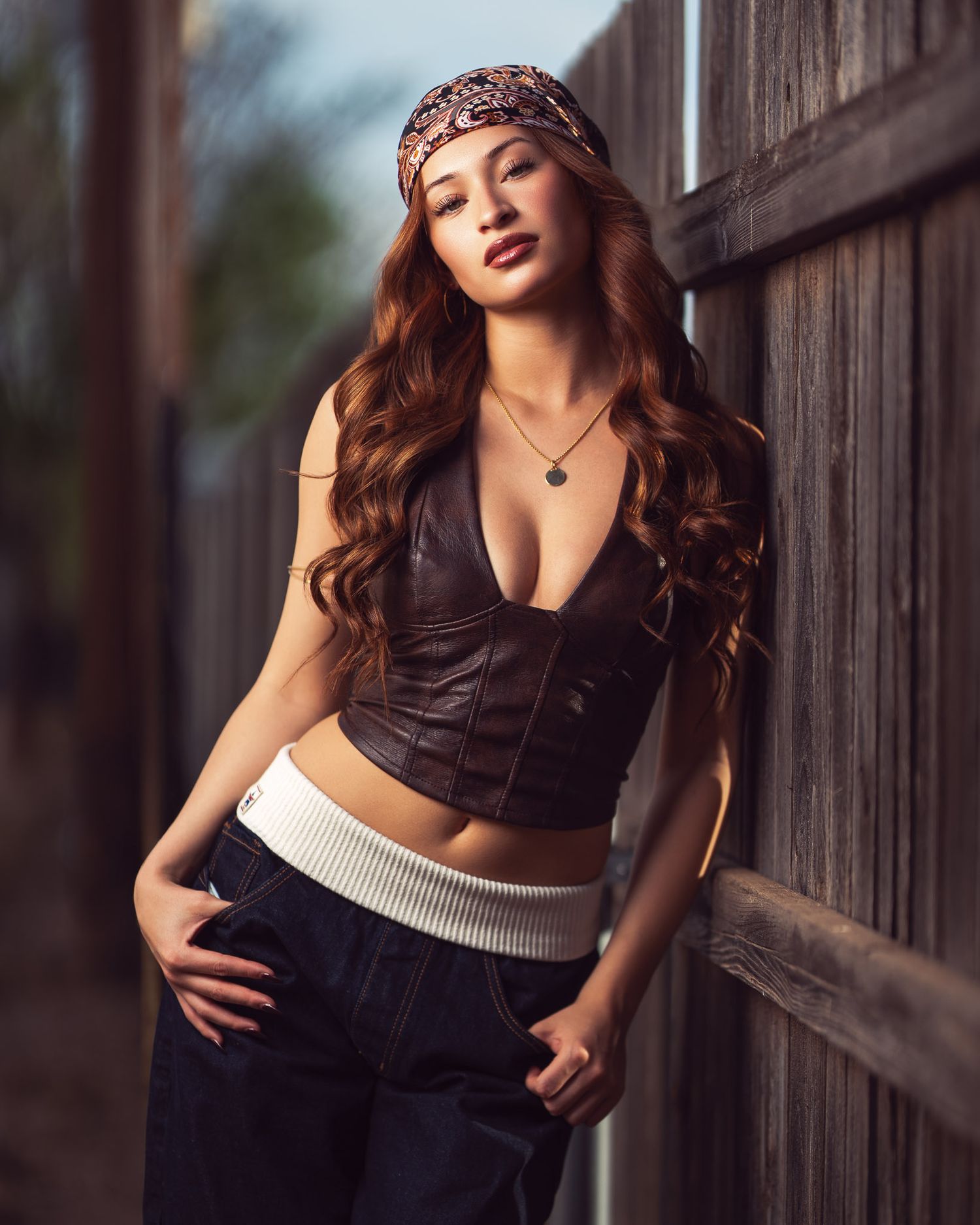 Young woman with long wavy auburn hair, a patterned bandana, and a brown leather corset top leans against a rustic wooden fence.
