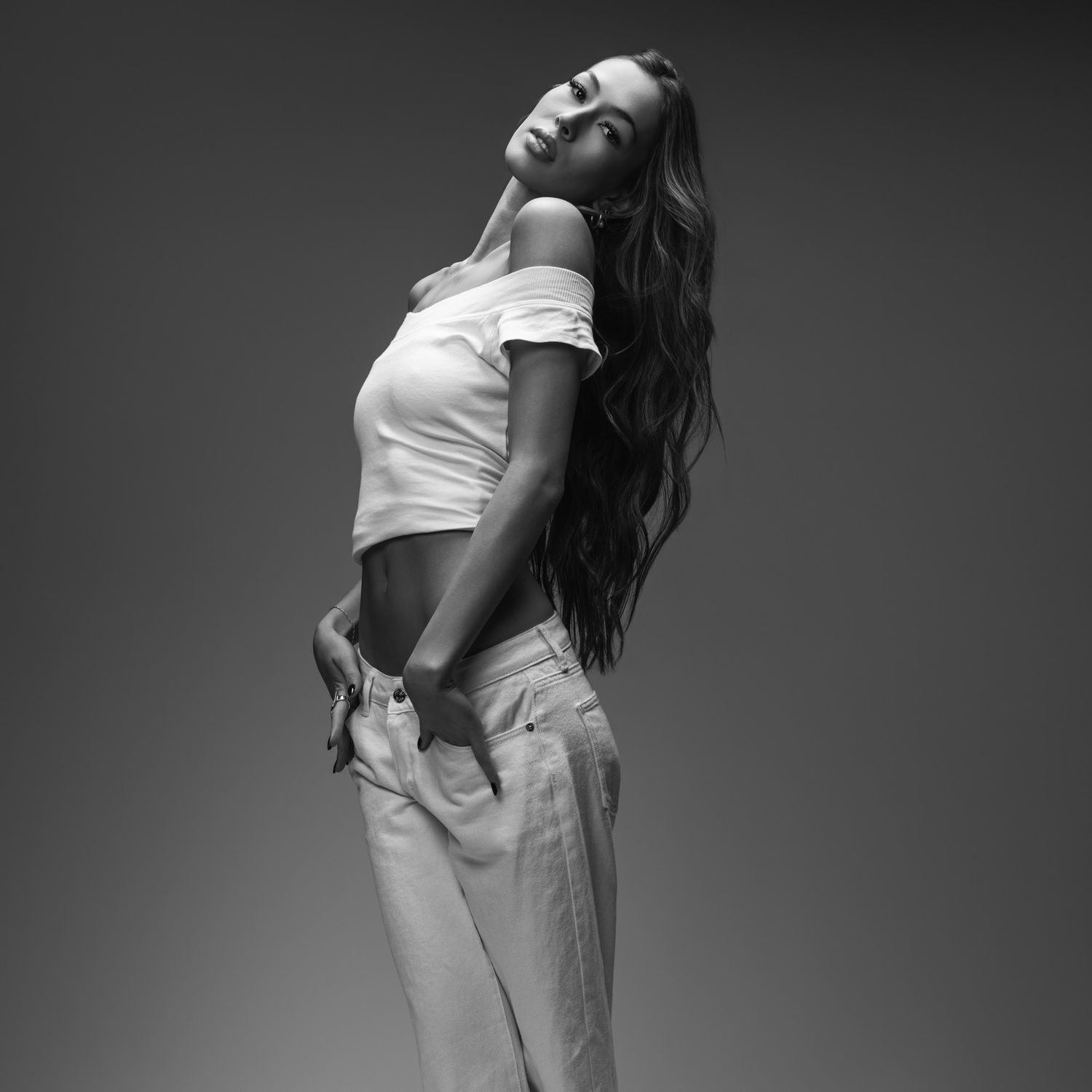 Black and white studio portrait of a woman with long wavy hair posing in an off-shoulder white crop top and loose light jeans.