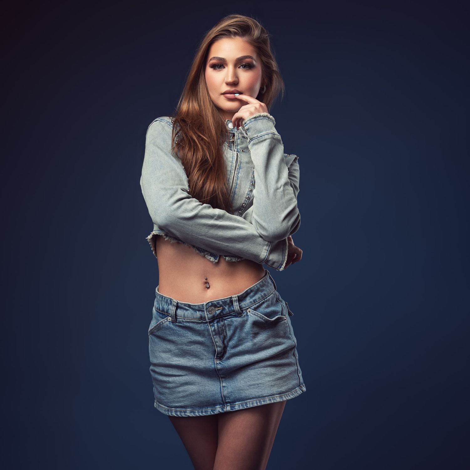Young woman with long brown hair poses in a gray cropped denim jacket and denim mini skirt against a dark blue studio background.