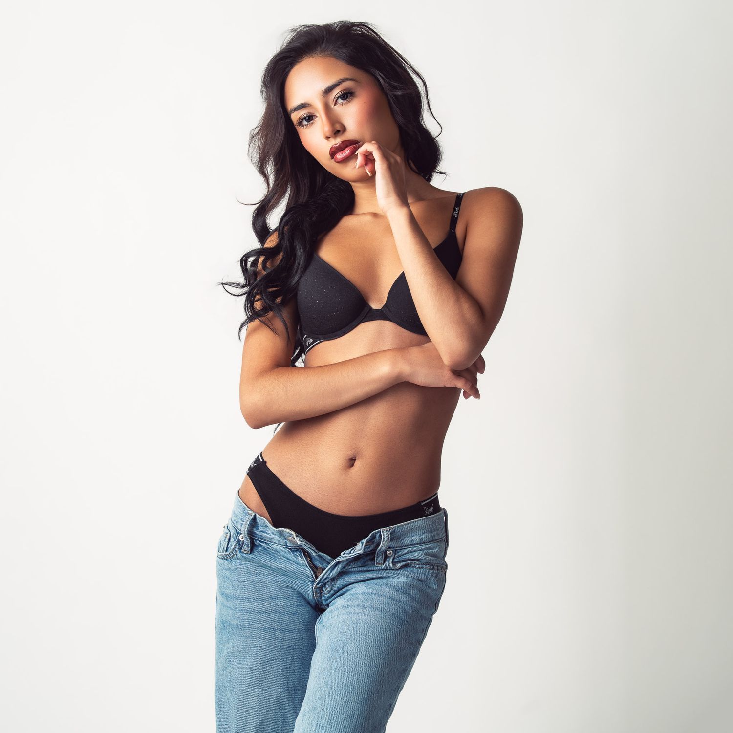 Woman with long wavy dark hair poses in a black bra and low-rise light wash jeans against a plain white studio background.