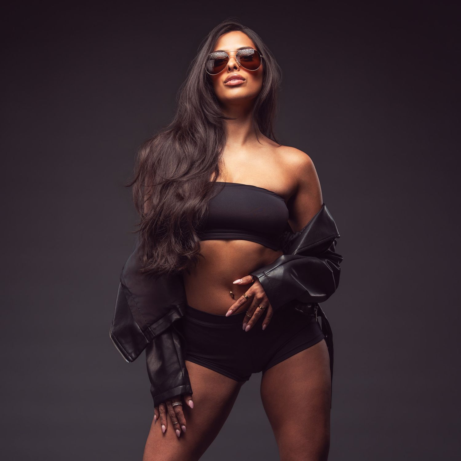 Woman with long wavy dark hair poses in sunglasses, a black bandeau top, shorts and an open leather jacket against a dark studio backdrop.