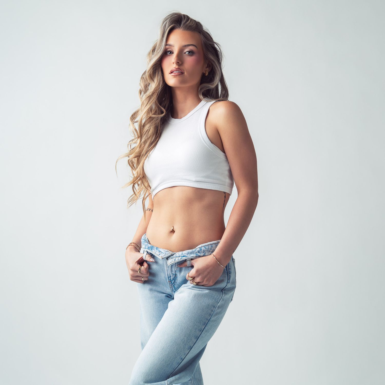 Woman with long wavy blonde hair poses in a white crop tank top and light-wash low-rise jeans against a plain light studio background.