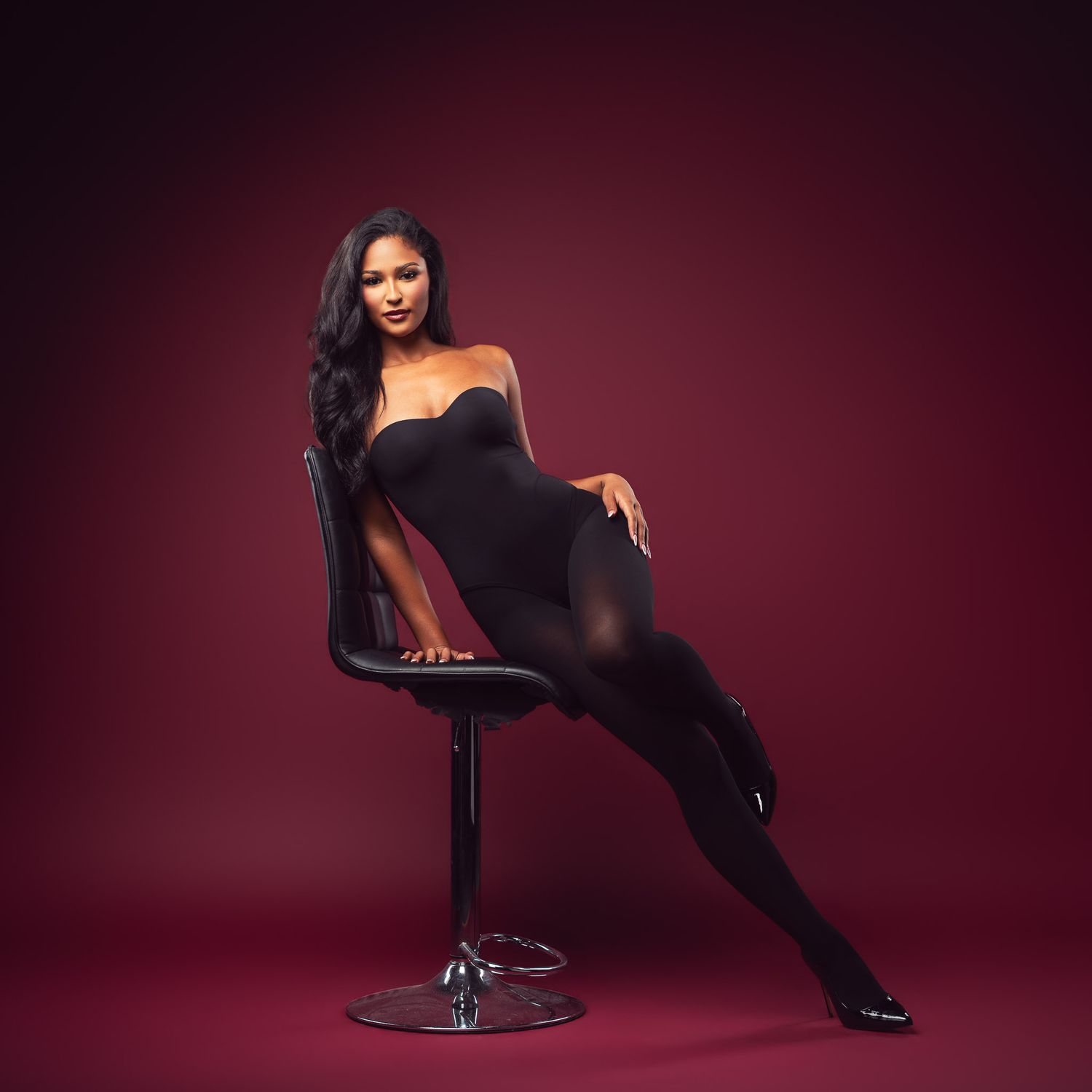 Woman with long dark wavy hair poses seated on a black swivel chair in a black strapless bodysuit against a deep burgundy studio backdrop.